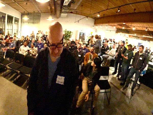 A wide-angle photograph captures a crowded indoor presentation or meetup in a loft-style space, showing a woman addressing an audience while a man in the immediate foreground, wearing a nametag, dominates the frame.