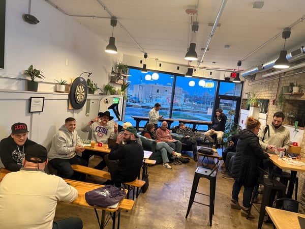 A group of people are socializing and relaxing in a casual indoor space that features a dartboard, an arcade game, and large windows looking out to a street at dusk.