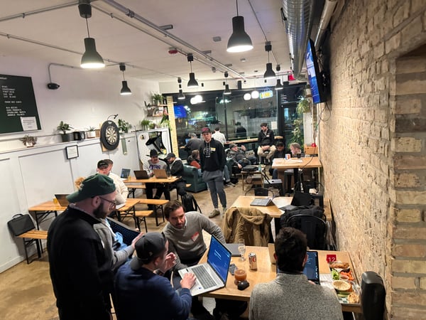 A group of people are gathered in a casual indoor space with brick walls, some working on laptops at wooden tables while others stand and talk.