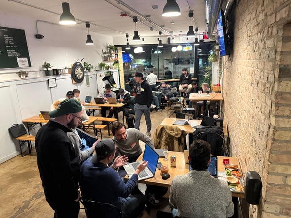 A group of people are gathered in a cafe or bar-like setting, many of whom are using laptops and engaged in discussions, suggesting a tech meetup or co-working event.