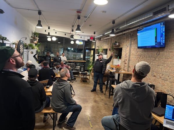 A group of people are gathered in an indoor space with brick walls, watching a presentation on a wall-mounted TV screen. A man stands at the front, pointing toward the screen as he speaks to the audience.