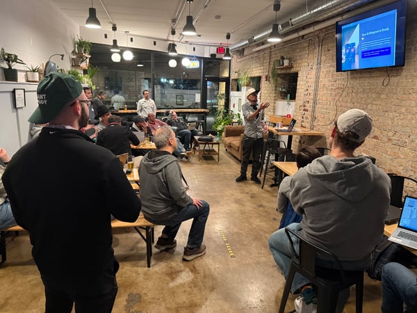 A man gives a presentation about an 'AI Wingman' app to a small audience in a room with exposed brick walls and a casual atmosphere.