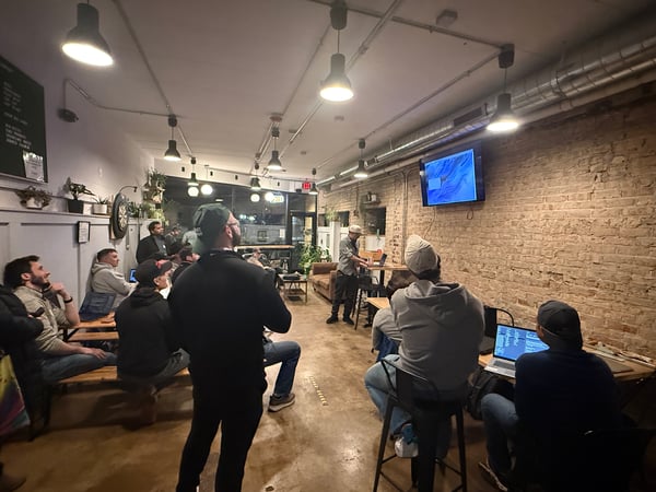A group of people are gathered in an indoor space with an exposed brick wall, watching a presentation or software demo on a wall-mounted screen.