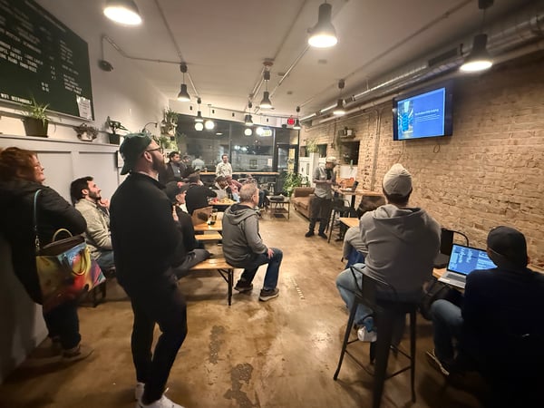 A group of people are gathered in a cozy, indoor space with exposed brick walls for a tech-related presentation.
