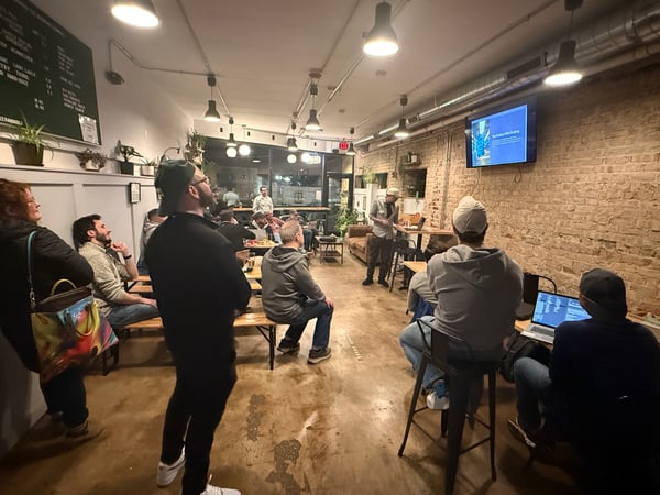 A group of people are gathered in a café or bar with exposed brick walls, watching a presentation titled 'The Problem with Budgets' on a wall-mounted screen.
