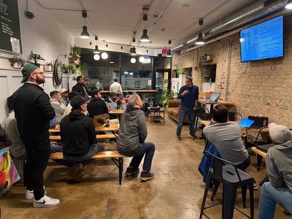 A group of people are gathered in an indoor space with exposed brick walls, watching a technical presentation where code is displayed on a large wall-mounted screen.