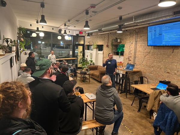 A man is giving a presentation to a group of people in an indoor space with exposed brick walls, while a large screen displays code.