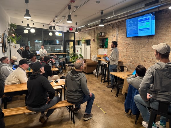A man is giving a presentation from a laptop to a seated audience in a room with exposed brick walls and industrial lighting.