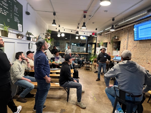 A group of people are gathered in an indoor space, likely a bar or cafe, watching a presentation being given by a man standing near a wall-mounted screen.