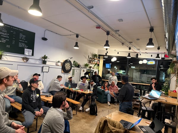 A group of people are gathered in a brewery taproom, listening to a man give a presentation. Several audience members have laptops open on tables in front of them.