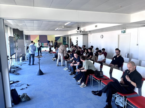 A group of people are gathered in a bright, modern office space for a presentation or meeting, with some sitting in rows and others standing near a whiteboard and screen.