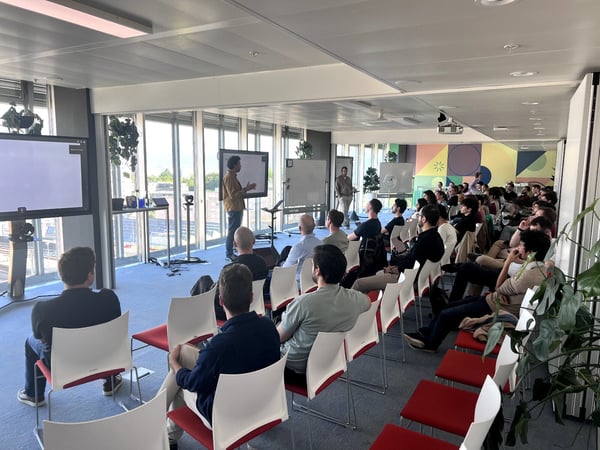 A man stands at the front of a modern, sunlit office space giving a presentation to a large audience seated in rows of chairs.