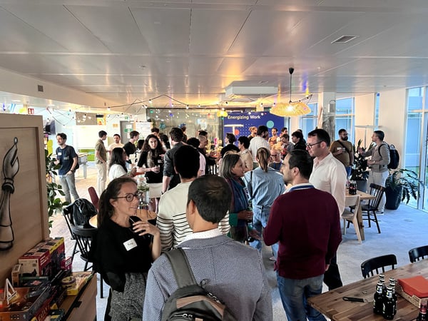 A large group of people are gathered in a modern, well-lit indoor office space for a social networking event, with board games visible in the foreground.
