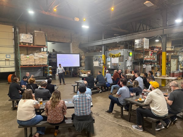 A man stands in front of a projector screen giving a presentation titled 'Reducing No Auth Claim Denials: Roadmap' to a group of people seated at picnic tables in a large warehouse-style venue.