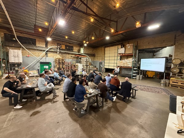 A group of people are seated at picnic tables in a large, industrial warehouse or brewery setting, attending a presentation shown on a large projector screen.