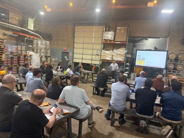 A group of people are seated at long tables in a brewery or warehouse setting, listening to a presentation displayed on a large screen.