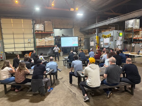 A man stands at the front of a large warehouse space giving a presentation to an audience seated at picnic tables.
