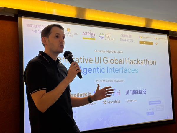A man is speaking into a microphone during a presentation in front of a large screen detailing a 'Generative UI Global Hackathon'.