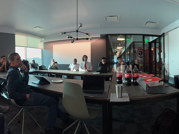 A group of people are gathered in a modern conference room for a presentation, with a man on the right gesturing towards a large screen showing 'Input Gateway'.
