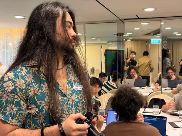 A man with long dark hair and a beard, wearing a tropical print shirt, stands in the foreground holding a camera. In the background, several people are seated at tables with laptops in a workshop or meetup setting.