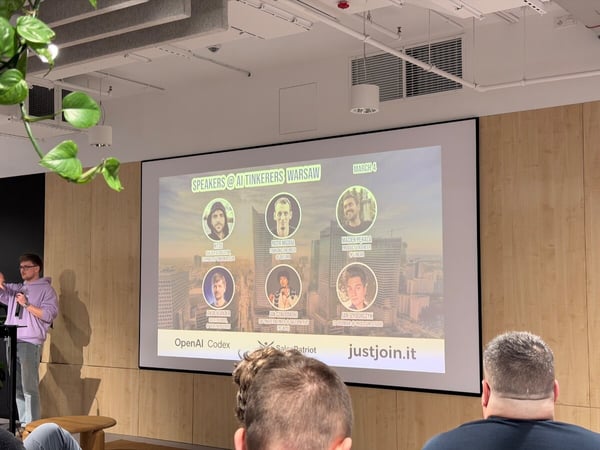 A man in a purple hoodie is giving a presentation at an 'AI Tinkerers Warsaw' event, standing next to a large screen displaying the names and photos of speakers.