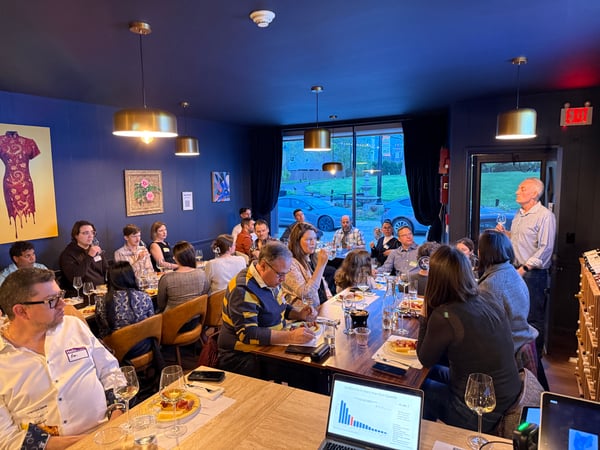 A group of people are gathered in a blue-walled room for a presentation and wine tasting event, with a speaker standing near a door.