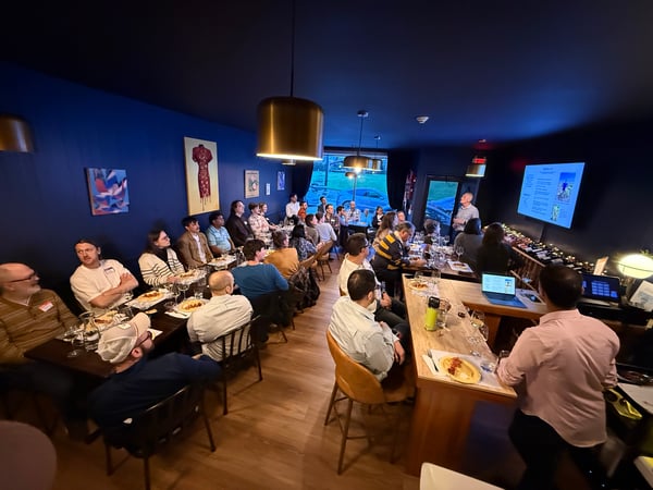 A group of people are gathered in a modern, blue-walled room for a presentation and dinner event. A presenter stands at the front near a screen while guests are seated at long wooden tables with food and drinks.
