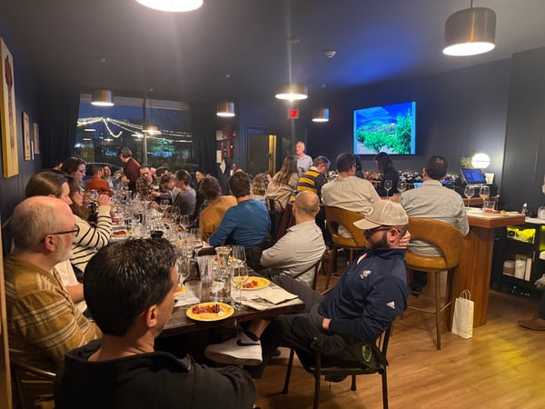A large group of people are gathered in a modern, dimly lit restaurant or event space, seated at long tables and a bar while a presentation is displayed on a screen.