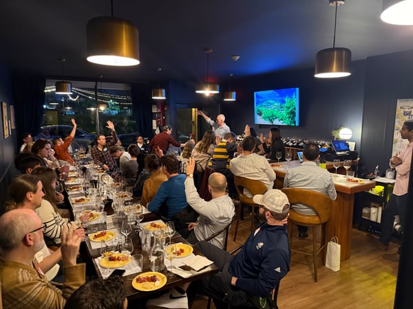 A group of people are gathered in a dimly lit restaurant or wine bar, many with their hands raised as they listen to a man giving a presentation near a TV screen.