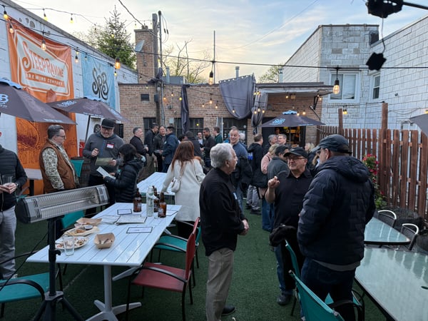 A group of people are gathered for a social event in an outdoor brewery patio area featuring string lights and banners.