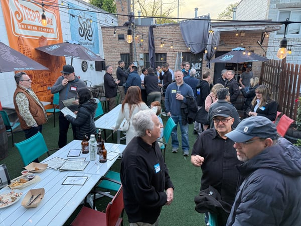 A group of people are socializing and networking at an outdoor brewery patio with tables, food, and drinks.