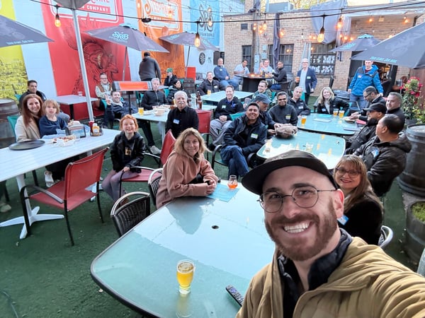 A large group of people are socialising at an outdoor brewery patio, with several seated at tables and a man in the foreground taking a selfie.