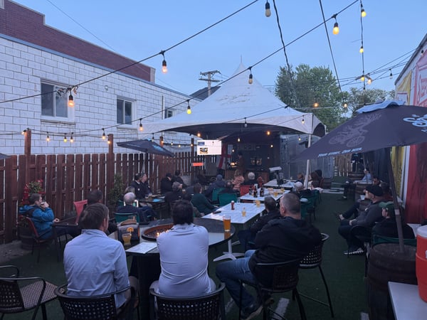 A crowd of people is gathered at an outdoor brewery patio during twilight, seated at tables and watching a presentation on a screen under a large white tent.