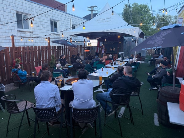 A group of people are gathered at an outdoor patio at dusk, many sitting at tables and watching a presentation being given under a white tent.