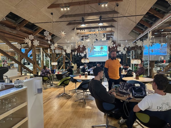 This is a candid photo taken inside a large, modern space featuring exposed wooden beams and winter snowflake decorations, where several people are gathered, including two men working on laptops in the foreground.
