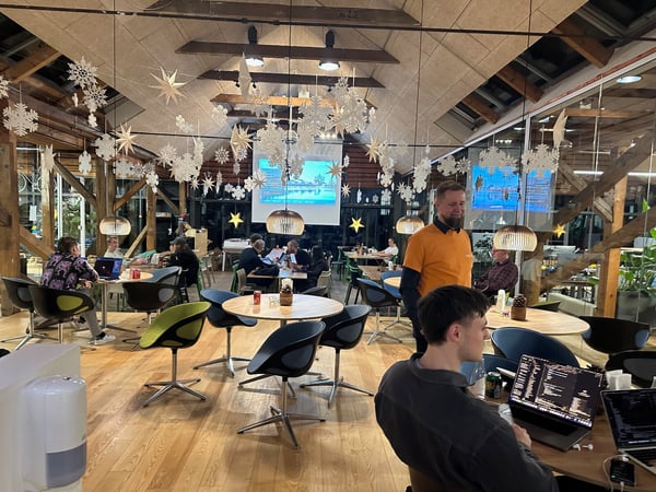 This is a candid photo of a group of people working or socializing in a large, warmly lit indoor space decorated for winter with hanging paper snowflakes and stars, featuring wooden beams and several individuals using laptops.