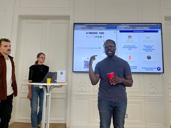 A photograph capturing an indoor event, likely a tech talk or presentation, where a speaker is gesturing toward a large digital screen listing the 'AI Tinkerers - Paris Main Stage Speakers' and their projects, with two other attendees standing nearby.