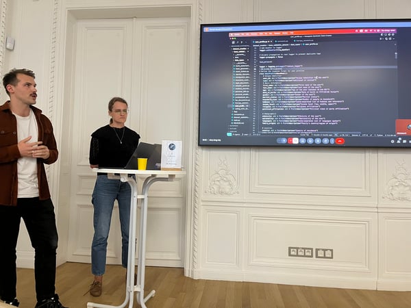 A photograph capturing a technical presentation indoors, featuring a man speaking and a woman standing behind a small podium with a laptop. A large screen behind them displays Python code defining data structures, set against an ornate white paneled wall.
