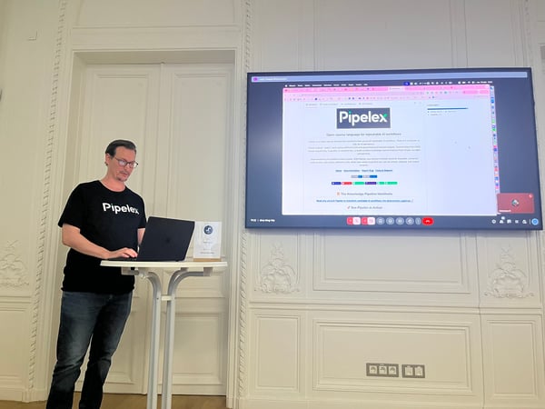 A man wearing a black 'Pipelex' t-shirt is giving a technical presentation, standing next to a laptop and podium, with his screen mirrored onto a large display showing the 'Pipelex' website in an ornate white room.