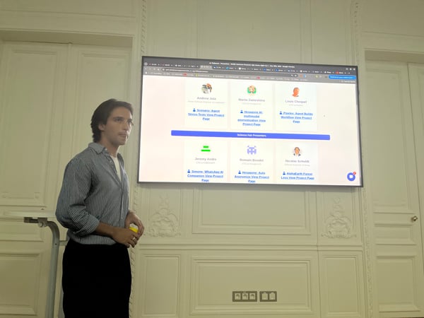 A young man stands next to a large wall-mounted screen displaying a list of 'Science Fair Presenters' and their AI-related projects in an ornate, white-paneled room.