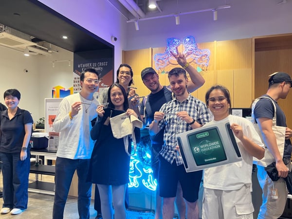 A group of eight people poses indoors at an event, smiling and holding up promotional items and a sign advertising the 'Lorong AI WORLDWIDE HACKATHON' sponsored by ElevenLabs.