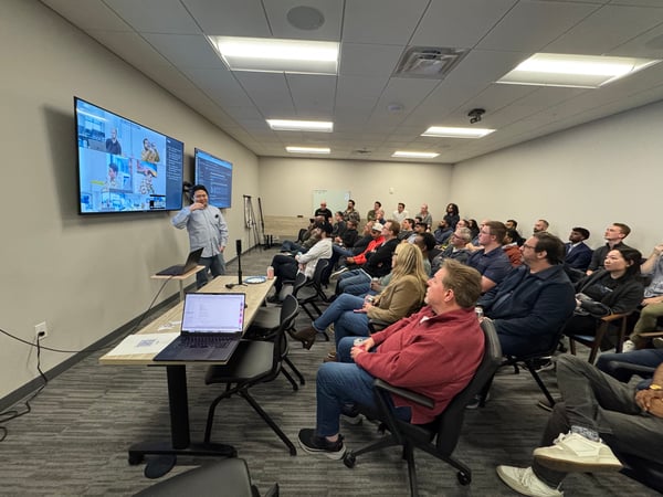 A man stands at the front of a conference room giving a presentation to a large seated audience, with two large screens displaying a video call and code.