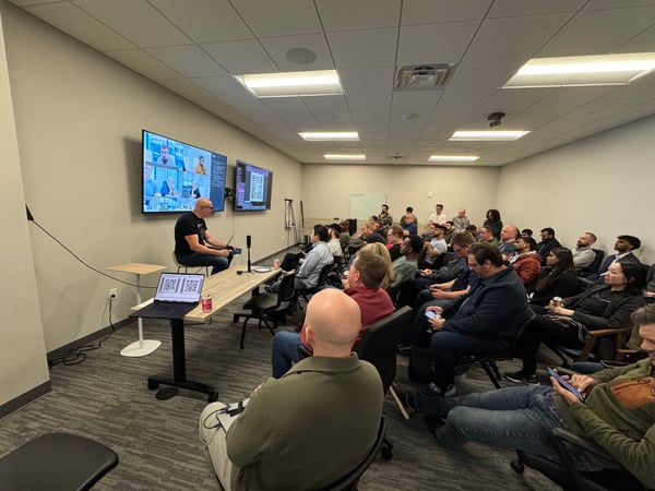 A group of people in a conference room attending a tech presentation, with a speaker seated at the front and screens displaying a video call and code.