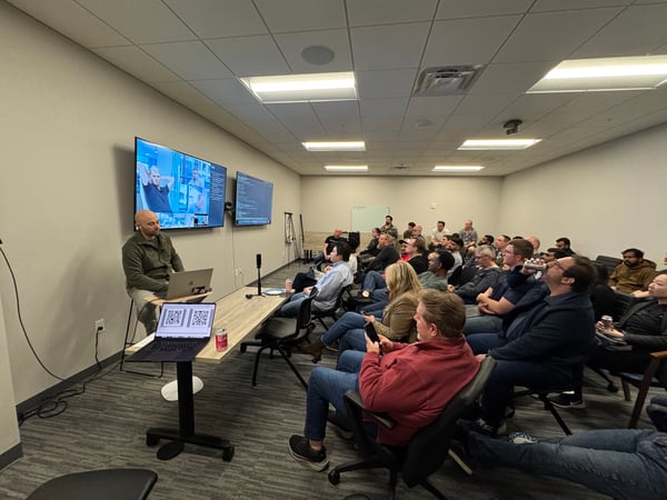 A man delivers a presentation to a group of people in an office setting, with two large screens displaying code and a video call.