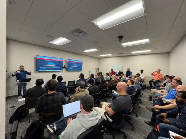 A man stands at the front of a conference room giving a presentation to a seated audience, with two large screens displaying technical diagrams.