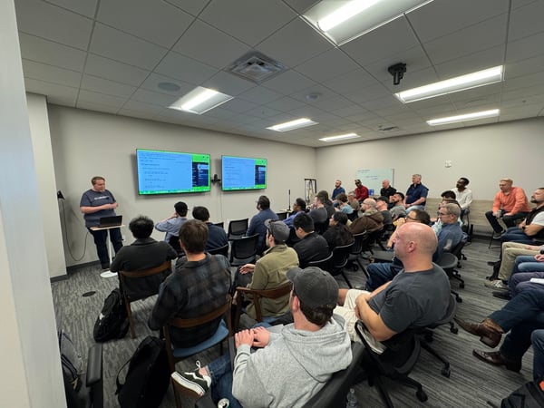 A man stands at the front of a room giving a presentation to a seated audience, with two large monitors displaying text behind him.