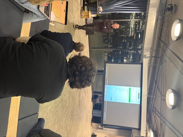 A man is giving a presentation in a modern indoor space at night, with audience members watching a screen displaying a software interface.