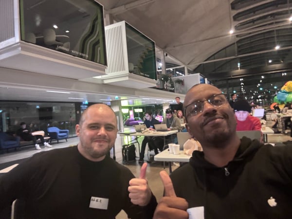 A selfie taken indoors at a large event or workspace, showing two men in the foreground smiling and giving thumbs-up gestures while many other participants work on laptops in the background.