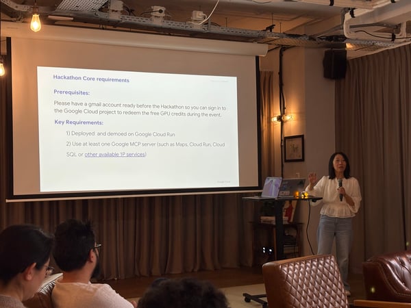 A woman stands beside a projector screen giving a presentation about Hackathon core requirements to an audience.