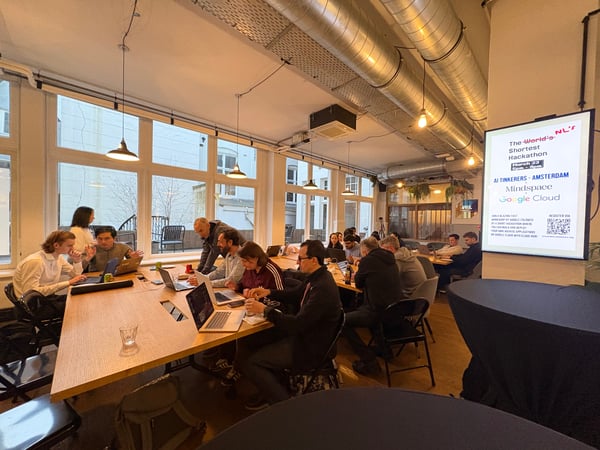 A group of people are gathered in a modern, industrial-style office space, working on laptops at long wooden tables during a hackathon event.
